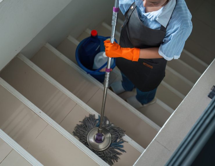 A cleaning woman is standing inside a building holding with a blue tank on the side facilities for tidying up in her hand.